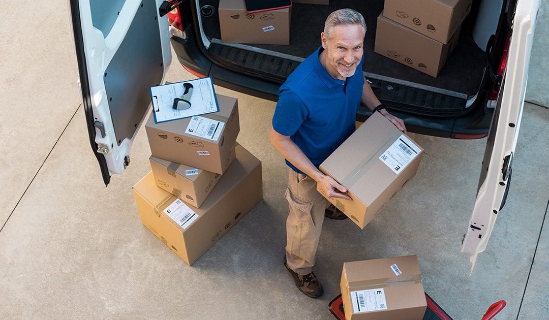 Happy delivery man holding cardboard package and looking at camera. Top view of courier loading packages in van for delivery. Portrait of happy man working at courier service and carrying parcels outside van.